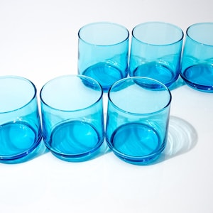 Könnte beinhalten: Sechs türkisfarbene Trinkgläser. Jedes Glas hat eine zylindrische Form mit abgerundetem Boden. Die Gläser sind in zwei Reihen auf einer weißen Oberfläche angeordnet, mit Schatten.