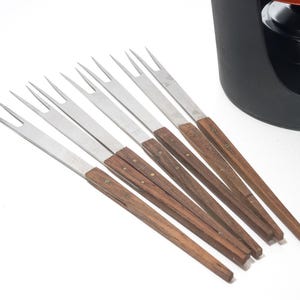 May include: A set of fondue forks with stainless steel tines and wooden handles. The forks are arranged on a white surface, with a black fondue pot in the background. The forks are approximately 8 inches long.