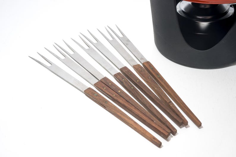 May include: A set of fondue forks with stainless steel tines and wooden handles. The forks are arranged on a white surface, with a black fondue pot in the background. The forks are approximately 8 inches long.