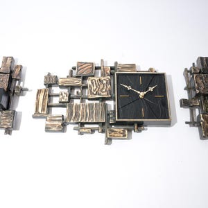 Könnte beinhalten: Ein Wanduhr-Set im Mid-Century-Modern-Stil mit einem quadratischen schwarzen Zifferblatt und goldenen Zeigern. Die Uhr ist von einem abstrakten Metalldesign mit zwei passenden Seitenteilen umgeben. Das Metall hat eine Bronze- und Silberoberfläche.