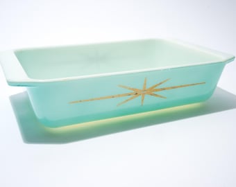 May include: A rectangular, turquoise baking dish with a white rim and a gold starburst design. The dish is empty and sits on a white surface, casting a shadow.