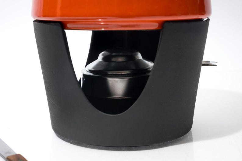 May include: Close-up of a fondue pot with a black metal stand and a bright orange pot. The stand has a curved design with an opening for the fuel source. The pot is round and sits securely on the stand.