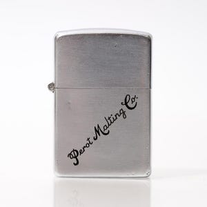 K&ouml;nnte beinhalten: Ein silberfarbenes Metallfeuerzeug mit dem Schriftzug "Perot Malting Co." in schwarzer Schrift. Das Feuerzeug hat eine rechteckige Form mit abgerundeten Kanten und einem Klappdeckel. Das Feuerzeug befindet sich auf wei&szlig;em Hintergrund.