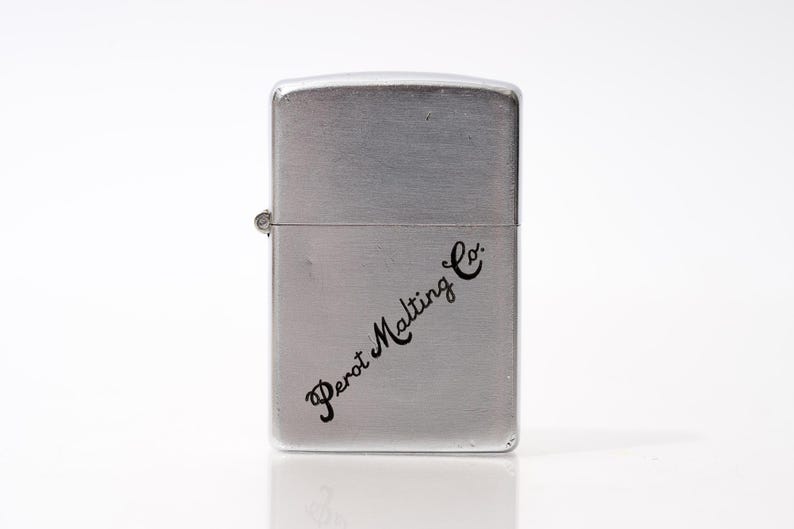 K&ouml;nnte beinhalten: Ein silberfarbenes Metallfeuerzeug mit dem Schriftzug "Perot Malting Co." in schwarzer Schrift. Das Feuerzeug hat eine rechteckige Form mit abgerundeten Kanten und einem Klappdeckel. Das Feuerzeug befindet sich auf wei&szlig;em Hintergrund.