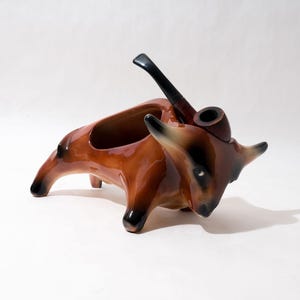 Könnte beinhalten: Eine braune Keramik-Stierfigur mit einer Pfeife im Maul. Der Stier dient als Aschenbecher und hat einen ausgehöhlten Rücken, um Zigarettenstummel aufzunehmen.