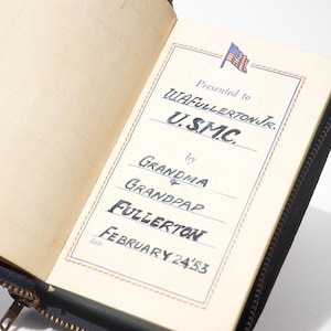 May include: Open vintage journal with a black cover and zipper. The cream-colored pages display handwritten text, including "Presented to W.A. FULLERTON JR. U.S.M.C." and "February 24 '53." A small American flag graphic is at the top.