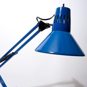 May include: A bright blue desk lamp featuring an adjustable articulated arm and a conical shade with vertical ventilation slits near the top. The lamp includes a black power cord and black mechanical joints connecting the metal arm segments.