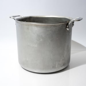 Puede incluir: Una olla de cocina grande, cilíndrica, de aluminio con dos asas curvas. La olla tiene una abertura ancha y un acabado mate ligeramente desgastado. Ideal para cocinar grandes cantidades de comida.