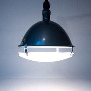 May include: A dark teal pendant light with a rounded shade and a white, curved diffuser. The light fixture is suspended from a black cord and emits a bright, diffused glow. The overall design is industrial.