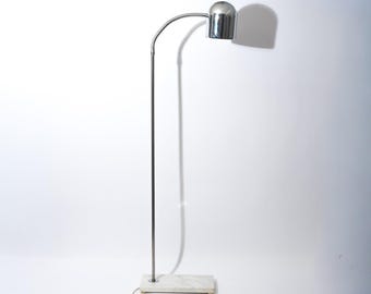 Pode incluir: Uma luminária de chão moderna com acabamento cromado. A luminária apresenta um braço flexível de pescoço de ganso e uma cabeça de lâmpada cilíndrica. A base é um bloco retangular de mármore branco. A luminária está ligada.
