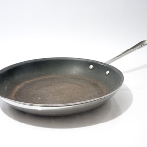 Könnte beinhalten: Eine gebrauchte, antihaftbeschichtete Bratpfanne mit einem langen, silbernen Griff. Das Innere der Pfanne ist dunkelgrau mit Gebrauchsspuren, während der Rand und der Griff silbern glänzen. Die Pfanne ist zum Kochen konzipiert.