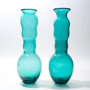 Peut inclure: Deux vases en verre turquoise avec une base bulbeuse et un col fin et ondulé. Les vases ont une finition lisse et brillante et sont présentés sur un fond blanc. Les vases mesurent environ 30 cm de haut.