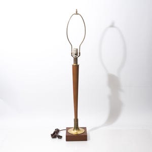 May include: A tall table lamp featuring a slender, tapered wooden body mounted on a square wooden base. The lamp includes brass-colored metal accents at the base and socket area, topped with a wire harp for a lampshade and a brown electrical cord.