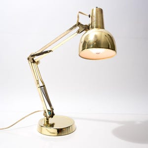 May include: A vintage brass desk lamp with an adjustable arm and a round base. The lamp has a classic design with a domed shade and a visible light bulb. The lamp is on a white surface.