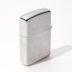 K&ouml;nnte beinhalten: Ein silberfarbenes Feuerzeug mit rechteckigem K&ouml;rper und abgerundeter Oberseite. Das Feuerzeug hat eine geb&uuml;rstete Metalloberfl&auml;che und einen Klappdeckel. Das Feuerzeug steht auf einer wei&szlig;en Oberfl&auml;che und wirft einen Schatten.