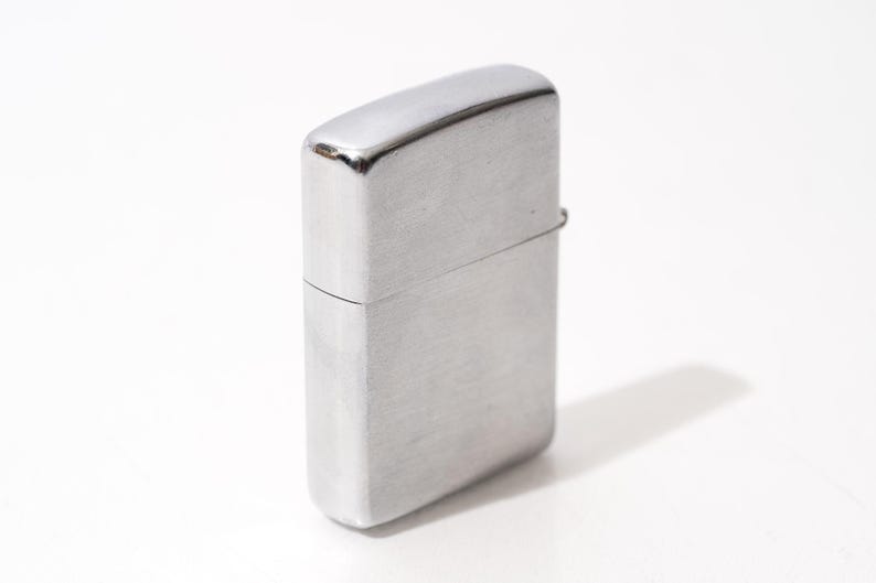 K&ouml;nnte beinhalten: Ein silberfarbenes Feuerzeug mit rechteckigem K&ouml;rper und abgerundeter Oberseite. Das Feuerzeug hat eine geb&uuml;rstete Metalloberfl&auml;che und einen Klappdeckel. Das Feuerzeug steht auf einer wei&szlig;en Oberfl&auml;che und wirft einen Schatten.