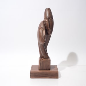 May include: A brown wooden sculpture of two intertwined faces, mounted on a square base. The sculpture has a smooth, polished finish and a minimalist design. The faces appear to be in a close embrace, creating a sense of intimacy.