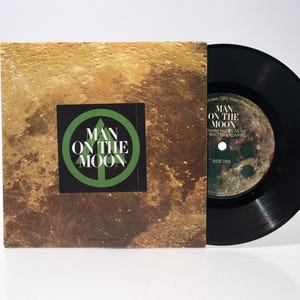 Könnte beinhalten: Eine Vintage 45 RPM Vinylplatte mit dem Albumcover für "Man on the Moon". Das Cover hat einen strukturierten, goldbraunen Hintergrund mit einem schwarzen Quadrat mit dem Text "Man on the Moon" in Grün. Die Schallplatte ist schwarz.