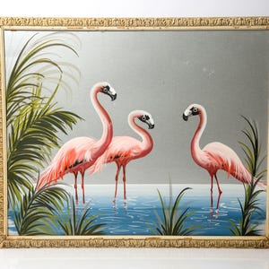 May include: A framed mirror featuring a painting of three pink flamingos standing in water. The artwork includes green foliage and is set against a gray background. The frame is gold-colored with ornate detailing.