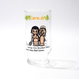 May include: Clear glass with a cartoon illustration of two figures, one holding a tennis racket. The glass has the words "love is" in green and a quote about tennis. A fun, novelty item.