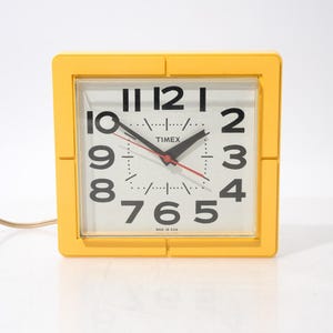 May include: A square electric wall clock with a bright yellow plastic frame and a white face. The clock features large black numerals, black hour and minute hands, a thin red second hand, and the Timex brand name printed in black text.