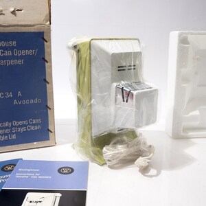 May include: A vintage Westinghouse Sesame Can Opener/Knife Sharpener, model HC 34 A, in Avocado color, is shown in its original packaging. The box and instruction manuals are visible, along with the can opener wrapped in plastic. The box reads "Hygienically Opens Cans, Can Opener Stays Clean, Re-usable Lid."