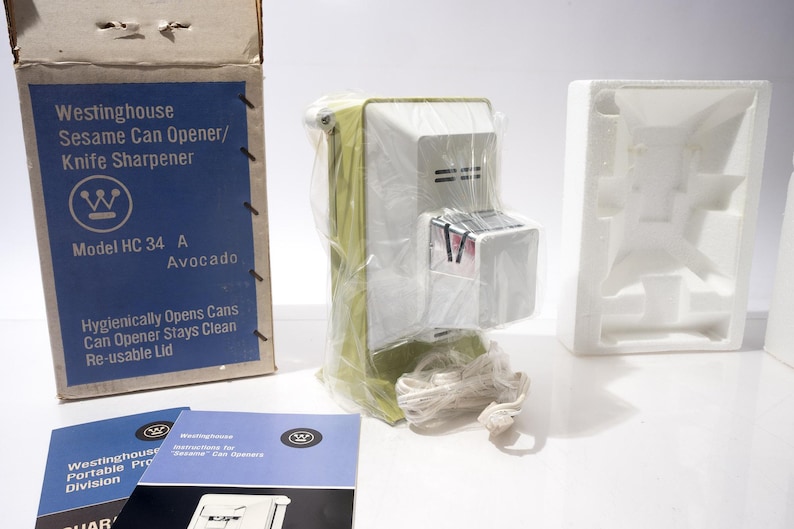 May include: A vintage Westinghouse Sesame Can Opener/Knife Sharpener, model HC 34 A, in Avocado color, is shown in its original packaging. The box and instruction manuals are visible, along with the can opener wrapped in plastic. The box reads "Hygienically Opens Cans, Can Opener Stays Clean, Re-usable Lid."