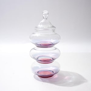 Puede incluir: Un dulcero de cristal transparente de tres niveles con tres secciones apiladas en forma de cuenco, con un sutil tono púrpura iridiscente y una tapa extraíble a juego rematada con un pequeño pomo redondeado como asa decorativa.