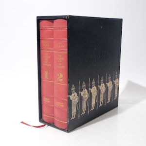 Op de afbeelding: Een set van twee rode hardcover boeken getiteld "The Ancient Art of Warfare" in een zwarte doos. De doos is versierd met goudkleurige illustraties van soldaten. Een rood lint-bladwijzer is zichtbaar.