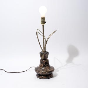 May include: A vintage table lamp with a bulb, featuring a dark brown base with gold accents. The lamp has a brass stem and leaf-like accents. The lamp is connected to a black power cord.