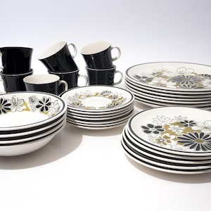 May include: A vintage dinnerware set featuring black mugs, bowls, plates, and saucers. The white plates and bowls have a black rim and a floral design in gold, black, and white. The mugs are solid black.