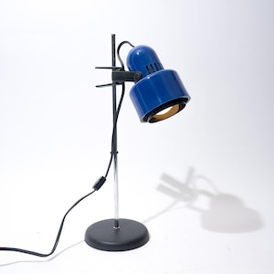 May include: A blue desk lamp with a cylindrical shade and a chrome stem. The lamp has a black base and a black power cord. The lamp is angled towards the viewer, and the light is on.