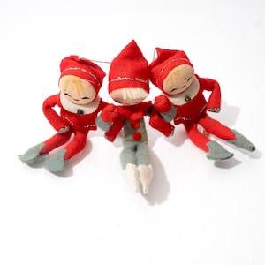 May include: Three small felt elf ornaments with painted faces and blonde hair. Two elves wear red outfits with silver trim and grey boots, while the center elf wears a red hat, a grey tunic with red buttons, and white boots. Each has a string for hanging.