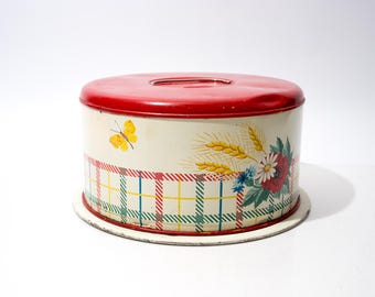 May include: A round metal cake storage tin with a bright red lid and a cream-coloured base. The side features a yellow butterfly, a cluster of wheat stalks, and a bouquet of red and white flowers, all set above a colourful plaid grid pattern.