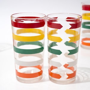 May include: Clear drinking glasses with horizontal bands of red, yellow, green, white, and orange. One glass has a zigzag pattern in the center. The glasses are arranged on a white surface.