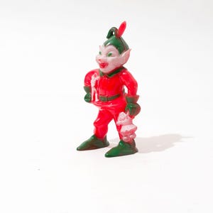 May include: A red and green plastic elf figurine with a pointed hat and a white face. The elf is holding a small white object in its hand.