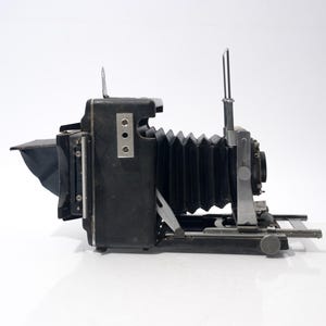 May include: A vintage black and silver large format camera with a bellows design. The camera has a lens, a viewfinder, and a sliding mechanism for focusing. It is set against a white background.
