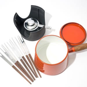 May include: An orange fondue pot with a wooden handle and lid, a black stand with a burner, and a set of fondue forks. The pot has a white interior. The forks have wooden handles and silver tines.
