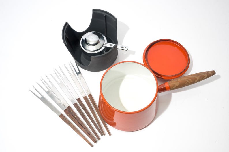 May include: An orange fondue pot with a wooden handle and lid, a black stand with a burner, and a set of fondue forks. The pot has a white interior. The forks have wooden handles and silver tines.