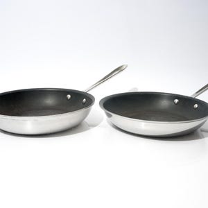 Può includere: Due padelle in acciaio inossidabile con interni neri e lunghi manici argentati. Le padelle sono esposte su una superficie bianca, mostrando il loro design elegante e moderno. Ideali per cucinare.