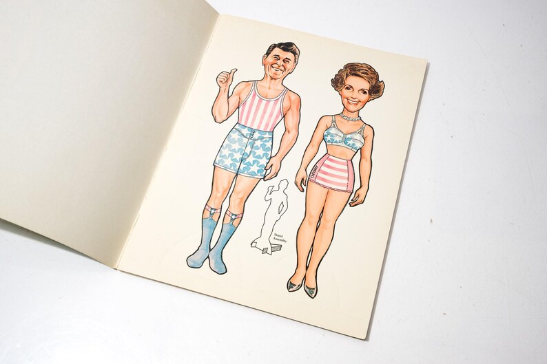 May include: Vintage paper dolls featuring a man in a striped tank top and star-patterned shorts, and a woman in a striped skirt and star-patterned bra. The illustrations are in color and are on a cream-colored paper.
