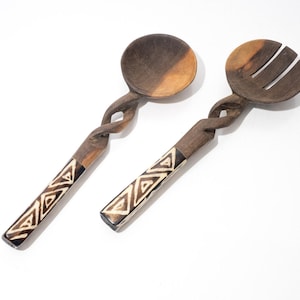 Może przedstawiać: Para drewnianych sztućców do sałatek. Jedna to łyżka, druga to widelec. Oba mają skręconą rączkę i geometryczny wzór w kolorze białym i czarnym. Łyżka ma 25 cm długości, a widelec ma 25 cm długości.