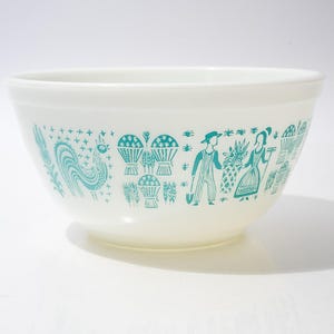 Puede incluir: Cuenco para mezclar de color crema con un diseño turquesa que representa un gallo, cestas y figuras en una escena rural. El cuenco tiene forma redondeada y probablemente está hecho de cerámica o vidrio.