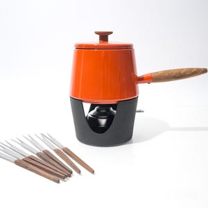Puede incluir: Una olla de fondue naranja con asa y pomo de madera, sobre una base negra. Varios tenedores de fondue con mangos de madera están dispuestos cerca. La olla es probablemente de metal esmaltado.