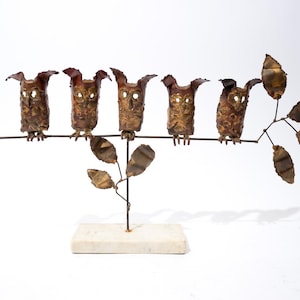 May include: A metal sculpture featuring six stylized owls perched on a horizontal branch with leaves. The owls and leaves have a bronze finish, and the sculpture is mounted on a rectangular, off-white base. The sculpture is a decorative item.