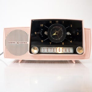 Può includere: Una radiosveglia vintage General Electric in plastica rosa con quadrante nero, numeri dorati, un orologio analogico circolare e una scala di frequenza radio orizzontale. L'unità include due manopole metalliche e una griglia per altoparlante.