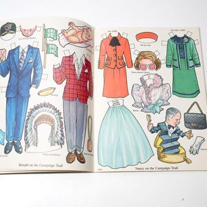 May include: Open book with vintage paper dolls. The left page features a blue suit, a red plaid jacket, and a Native American headdress. The right page shows a red suit, a green suit, and a light blue skirt. Text reads "Ronald on the Campaign Trail" and "Nancy on the Campaign Trail."