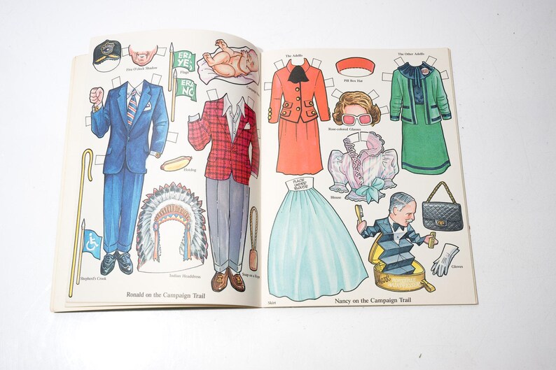 May include: Open book with vintage paper dolls. The left page features a blue suit, a red plaid jacket, and a Native American headdress. The right page shows a red suit, a green suit, and a light blue skirt. Text reads "Ronald on the Campaign Trail" and "Nancy on the Campaign Trail."
