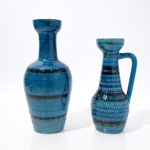 May include: Two blue ceramic vases with a glossy finish. The taller vase has a wide neck and bulbous body. The shorter vase features a single handle and a tapered neck. Both pieces are decorated with horizontal bands of dark blue patterns.