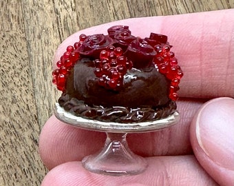 May include: A miniature chocolate cake decorated with red candy roses and red candy beads on a clear cake stand.